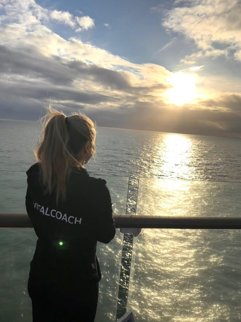 Frau schaut von Kreuzfahrt Schiff auf Meer. Auf dem Rücken steht Vitalcoach für Ayurveda Massagen